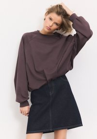 Giovane donna che indossa una felpa viola larga e una gonna di jeans scura, che posa con una mano nei capelli biondi davanti a uno sfondo neutro.