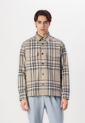 Beige plaid shirt made of fabric with a soft texture; features dark blue and black lines, two chest pockets, and a button-up front.
