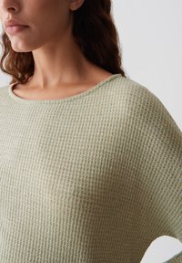 Femme aux longs cheveux bruns portant un pull en tricot texturé vert clair à encolure ronde sur un fond uni.