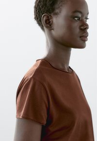 Brun crew-neck t-shirt med korte ærmer, lavet af glat stof. Nærbillede af en person iført t-shirten, med naturlig makeup.