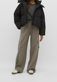 Sort pufferjakke med tyk polstring, grå sweatshirt under, og brede olivengrønne cargo bukser med sidelommer. Hvide sneakers synlige.