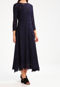 Granatowa koronkowa sukienka z długimi rękawami i pełną spódnicą, z kwiatowymi wzorami i falbaniastym wykończeniem. Noszona z czarnymi szpilkami na kostkę.