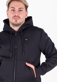 Felpa nera con zip, cappuccio, tasche frontali e cordini regolabili. Presenta una toppa con logo sulla manica e un tessuto nero strutturato.