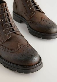 Brune læder snørebåndsstøvler med broguedetaljer, rund tå og en robust sort gummisål. Tekstureret finish med synlige sømme.