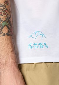 Weißes Baumwoll-T-Shirt mit einem blauen Bergumriss und geografischen Koordinaten nahe dem unteren Saum. Sichtbarer Hautton und Textur.
