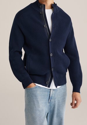 Hombre con cárdigan azul marino acanalado con cremallera y botones sobre camisa blanca y vaqueros azul claro, con las manos relajadas a los lados.