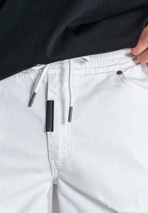 Person trägt weiße Hose mit Kordelzug und ein schwarzes Hemd, hebt mit einer Hand das Hemd an, um den Bund und den Kordelzug zu zeigen.