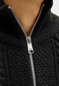 Czarny sweter dzianinowy z wysokim kołnierzem, z metalowym zamkiem błyskawicznym i teksturowanym materiałem. Na suwaku znajduje się grawerowane logo marki.