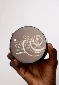 Contenant rond noir étiqueté « Patch de gel pour les yeux intensif à la réparation des escargots en or » avec texte et logo blancs ; finition lisse, tenu dans une main.