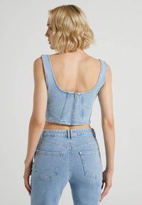 Top in denim azzurro chiaro, corto, con spalline larghe, schiena a scoperta e chiusura con cerniera sul retro. Presenta un design aderente e una morbida texture.