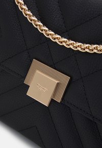 Czarna pikowana torebka z fakturowanej skóry, złotym łańcuszkiem jako paskiem i prostokątnym zapięciem z pionowymi liniami oraz logo.