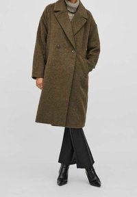 Zelený strukturovaný dvouřadý kabát s velkými klopami, dvěma knoflíky, bočními kapsami a volným střihem, nošený přes pletený svetr a černé kalhoty.