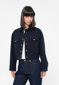 Navy cropped jacket in textured fabric with gold buttons, two chest pockets, and a shirt collar, paired with dark denim jeans.
