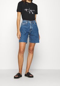 Denim shorts with a high waist, blue color, frayed hem, paired with a black t-shirt featuring white Calvin Klein logo, and black slide sandals.