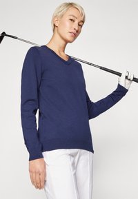 Un pull-over en v bleu marine fabriqué en tissu lisse, avec des manches longues et une taille côtelée ajustée. Un club de golf est posé sur l'épaule.