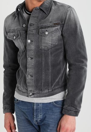 Mand iført en falmet sort denimjakke knappet over en grå skjorte og blå jeans mod en ensfarvet baggrund.