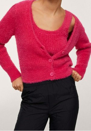 Personne portant un cardigan duveteux rose vif par-dessus un débardeur rose assorti, accompagné d'un pantalon noir ample, sur un fond uni.