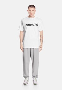 Mężczyzna stojący przodem, ubrany w białą koszulkę PRNTN, jasnoszare spodnie dresowe, białe i zielone sneakersy oraz z tatuażami na lewej ręce i dłoniach.