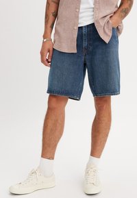 Dunkelblaue Denim-Shorts mit lockerer Passform, ausgestattet mit Gürtelschlaufen, Seitentaschen und sichtbaren Nähten. Kombiniert mit weißen Sneakers und Socken.