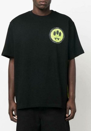 Mies, joka käyttää löysää mustaa t-paitaa, jossa on pieni keltavihreä ympyräembleemi, jossa on kolme mustaa silmää ja kaksi fangsia ylävasemmalla rinnassa.