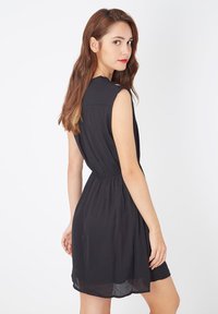 Joven mujer con cabello largo y castaño, vistiendo un vestido negro sin mangas, de pie de lado y mirando por encima del hombro contra un fondo blanco.