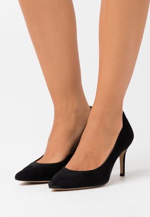 Woman wearing black suede pointed-toe high heels with mid-height stiletto heels on a white background.