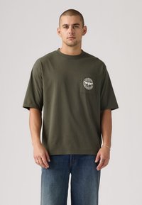 Χακί βαμβακερό t-shirt με κοντά μανίκια, στρογγυλή λαιμόκοψη και τσέπη στο στήθος με κυκλικό γραφικό σχέδιο.