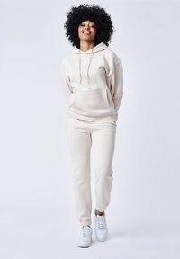 Sudadera beige con bolsillo frontal y cordón, combinada con joggers a juego. Ambas prendas tienen una textura suave y puños elásticos, llevadas con zapatillas blancas.