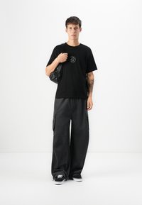 Junger Mann in schwarzem Grafik-T-Shirt, weiten dunklen Cargo-Hosen, schwarzen Sneakers und mit einer schwarzen Umhängetasche, der vor einem weißen Hintergrund steht.