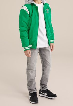Chaqueta bomber acolchada verde con capucha gris, acentos en los hombros blancos y puños a rayas. Llevada sobre una camiseta blanca y combinada con jeans grises y zapatillas negras.
