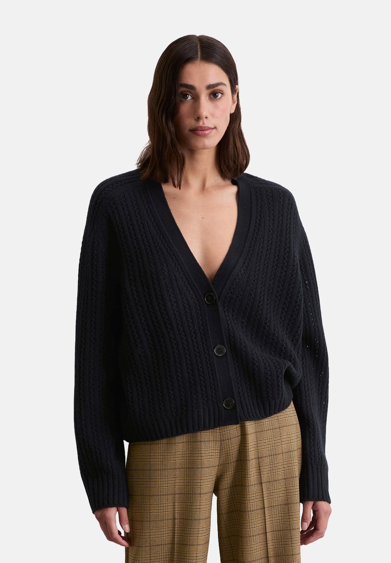 Schwarze Strickjacke mit tiefem V-Ausschnitt und fünf Knöpfen, mit strukturierter Rippung und lockerer Passform, kombiniert mit karierten Hosen.