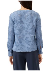Lichtblauwe blouse met een textuur van bladeren, doorschijnende stof, knoopmanchetten en rafelige details langs de naden en randen.