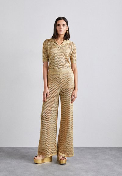 Ensemble en tricot composé de deux pièces en or avec un motif en chevrons ; top à col à manches courtes, pantalon à jambes larges et chaussures à talons compensés dorées.
