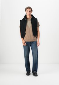 Joven de pie, vistiendo una camiseta gráfica marrón, un suéter negro sobre los hombros, jeans azules y zapatos negros sobre fondo blanco.