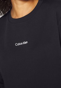 Camiseta de algodón negra con cuello redondo y el logo blanco de "Calvin Klein" en una fuente sans-serif moderna, con una textura suave.