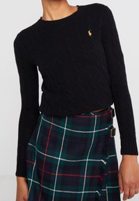 Svart kabelstickad tröja med rund halsringning och långa ärmar, med en liten guldig broderad logotyp på bröstet. Kombinerad med en rutig kjol.