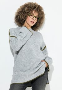 Szary, oversize sweter z zielonym wykończeniem, z prążkowanymi mankietami i bocznymi rozporkami. Model nosi duże prostokątne zielone okulary.