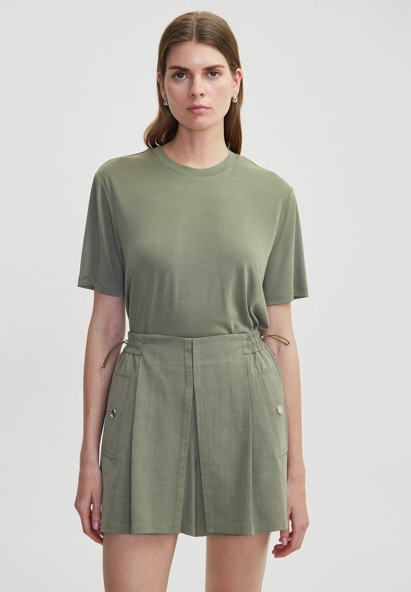 Camiseta de manga corta color verde oliva con escote redondo, combinada con pantalones cortos de cintura alta que presentan pliegues y bolsillos laterales, todo en un tono a juego.