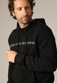 Zwarte hoodie van katoen, met een voorste kangaroozak, een capuchon met trekkoord en "DEELUXE ORIGINAL" in het wit gedrukt op de borst.