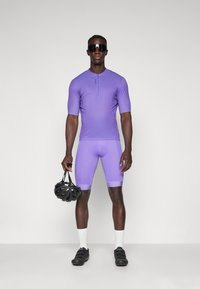 Lilla sykkeldrakt med korte ermer og glidelås halvveis oppe. Sykklisten har på seg matchende lilla shorts, svarte sko og holder en svart hjelm.