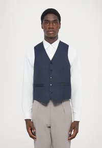 Gilet en tissu bleu marine avec une coupe ajustée, cinq boutons noirs à l'avant, deux poches et un ourlet pointu, porté par-dessus une chemise blanche.