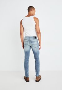 Jeans azules desvaídos con un ajuste slim, que presentan costuras en contraste, cinco bolsillos y una etiqueta negra en la cinturilla. Llevados con una camiseta sin mangas blanca.