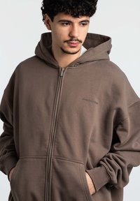 Felpa con cerniera marrone, dotata di un ampio cappuccio, tasche frontali e polsini a costine. Realizzata in un tessuto morbido con un discreto dettaglio del logo.