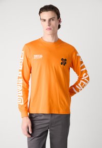 Narančasta majica dugih rukava od pamuka s crnim cvjetnim uzorkom na prsima i bijelim grafičkim tekstom duž rukava.