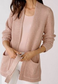 Cárdigan rosa claro de punto con mangas anchas, dos bolsillos delanteros y un ajuste holgado. La tela texturizada añade dimensión.