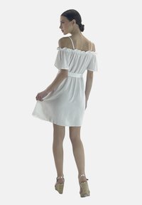 Vestido blanco off-shoulder con detalle de volantes, cinta en la cintura y falda acampanada. La modelo lleva tacones de cuña. La tela parece ligera y fluida.