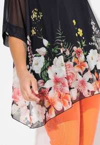 Top nero trasparente con motivo floreale in rosa e bianco, caratterizzato da una vestibilità ampia e maniche corte, abbinato a pantaloni arancioni brillanti.