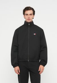 Sort zip-up jakke med høj krave, elastiske manchetter og bund. Har et lille flerfarvet logo på venstre bryst. Glat stofstruktur.