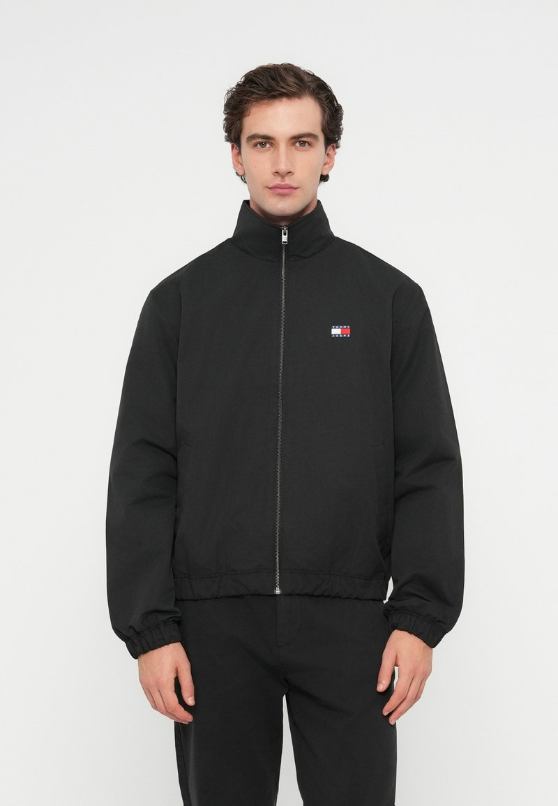 Sort zip-up jakke med høj krave, elastiske manchetter og bund. Har et lille flerfarvet logo på venstre bryst. Glat stofstruktur.