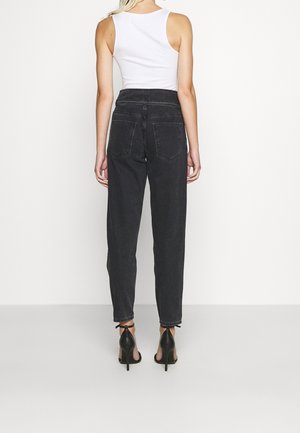 Kvinde, der står med ryggen til iført hvid ærmeløs top, sorte høj taljede jeans og sorte høje hæle sandaler mod en ensfarvet baggrund.